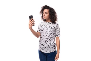 cute curly young brunette with shoulder-length hair dressed in a short-sleeved shirt makes a selfie on the phone.