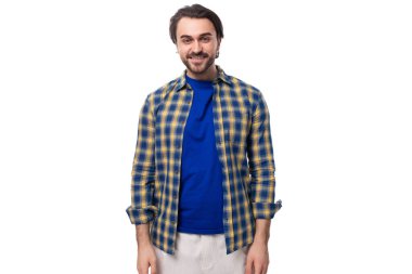 Sakallı, küpeli, beyaz bir arka planda mavi ekose gömlek ve tişört giyen beyaz saçlı esmer bir adam..