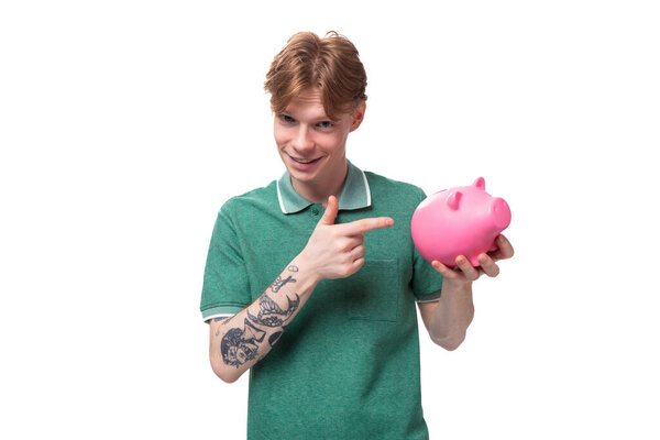a young European man with red hair holds out a pink piggy bank with money and coins.