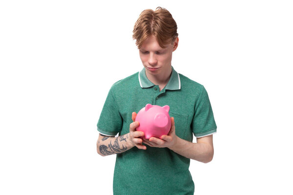 a young student of a red-haired man in a green t-shirt with a tattoo on his arm accumulated money in a piggy bank.