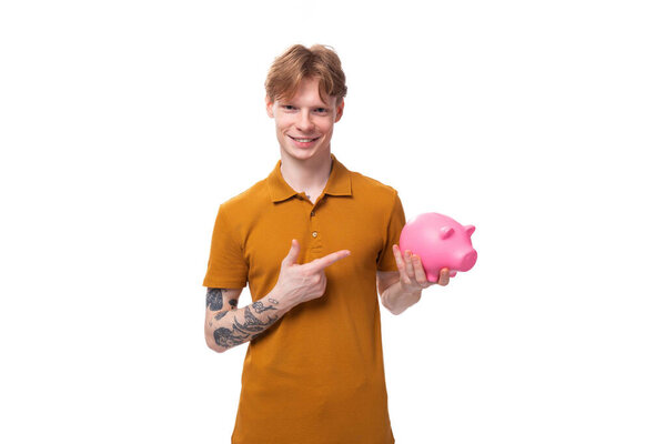 a young successful red-haired guy dressed in an orange t-shirt with a tattoo on his arm holds savings in a piggy bank.
