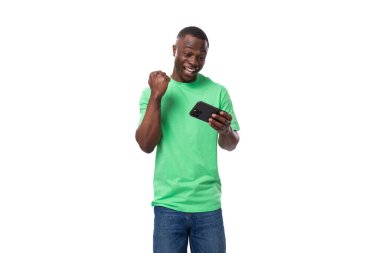 Açık yeşil tişörtlü ve kot pantolonlu Afrikalı genç adam akıllı telefondan video izliyor..