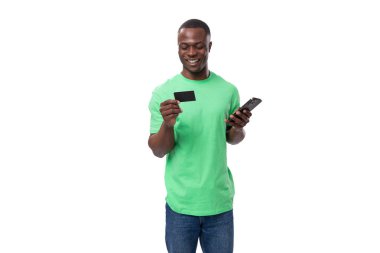 Yeşil tişört ve kot pantolon giyen genç, mutlu, dost canlısı Amerikalı adam cep telefonu ve kredi kartı kullanıyor..
