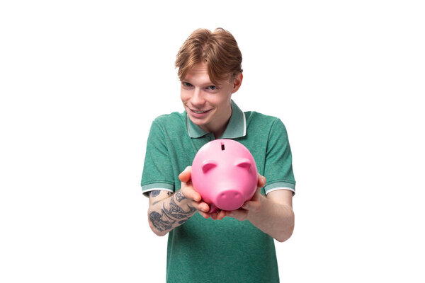 a young European man with red hair holds out a pink piggy bank with money and coins.