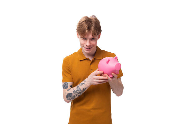 young red-haired guy in a yellow t-shirt holding a piggy bank on a white background with copy space.