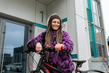 Bisiklet kiralama konsepti. Mor kürklü rastalı genç bir kadın bisiklet sürüyor..