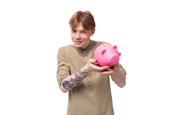 young red-haired guy in glasses and a brown shirt holds a pink piggy bank.