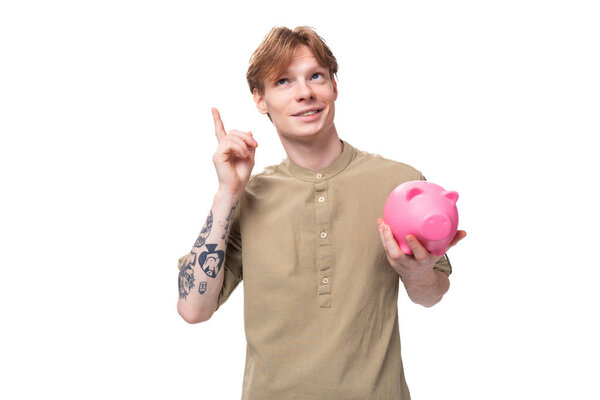 young handsome red-haired man in glasses and a shirt keeps savings in a piggy bank on a white background.