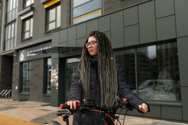 Sokakta çekilmiş bir fotoğraf. Saçları dağınık, ceketli, bisiklete binen genç aktif bir kadın..