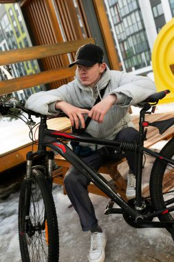 Şapkalı ve ceketli bir adam bankta bisikletle dinlenmek için oturdu..