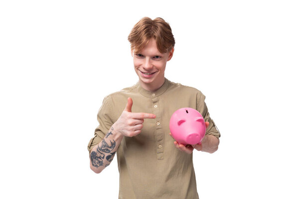 young handsome red-haired man in glasses and a shirt keeps savings in a piggy bank on a white background.