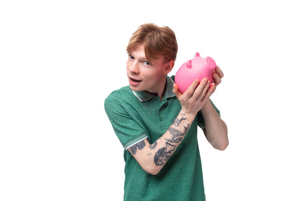 joyful happy young red-haired guy in a green t-shirt holds a piggy bank with money and thinks where to spend.