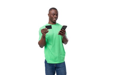 Yeşil tişört ve kot giyen genç Amerikalı adam akıllı telefon ve kredi kartı kullanıyor..