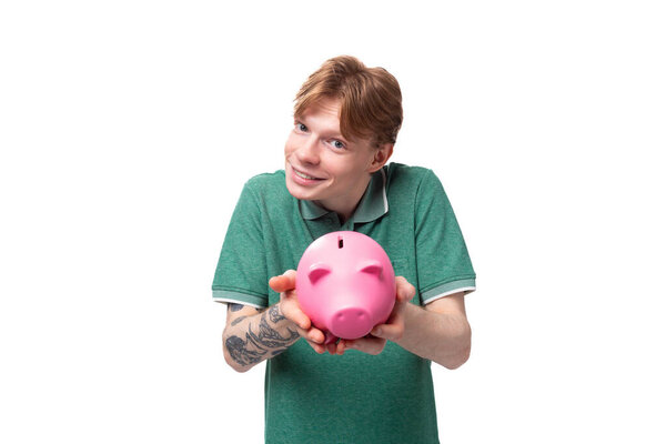 a young student of a red-haired man in a green t-shirt with a tattoo on his arm accumulated money in a piggy bank.