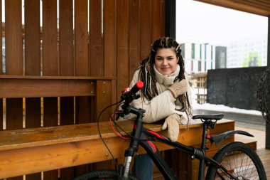 Bir kadın, bisikletin yanındaki ahşap bir bankta oturuyor. Rahatlamış görünüyor ve günlük giysiler giyiyor. Bisiklet bankın karşısına park edilmiş, bu da bisiklette mola ya da duraklama olduğunu gösteriyor.