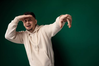 Kapüşonlu bir adam elleri başının arkasında duruyor, rahat ve kendinden emin görünüyor. Arka plan basit ve tanımsız, erkeklerin duruş ve ifadelerine dikkat çekiyor..