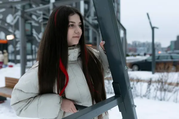 A woman with long brown hair is standing in the snow, surrounded by a white winter landscape. She is looking ahead, bundled up in warm clothing to protect against the cold weather.