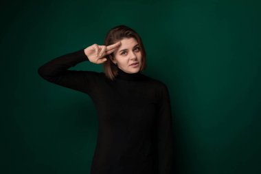 Siyah gömlek giyen bir kadın fotoğraf için poz veriyor. Elleri kalçasında kameraya gülümserken kendine güvenerek duruyor. Arkaplan basit, odak noktasının sadece