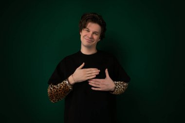 Sıradan kıyafetler giyen bir adam katı yeşil bir duvarın önünde duruyor. Rahatlamış görünüyor ve ileriye bakıyor. Duvar, erkeğin varlığı için basit bir arkaplan sağlıyor..