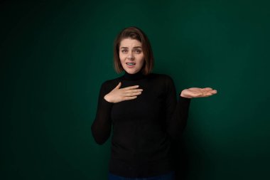 Canlı yeşil bir duvarın önünde dimdik duran bir kadın. Doğrudan kameraya tarafsız bir ifadeyle bakıyor gibi görünüyor. Arkaplan koyu yeşil ve görünür bir deseni yok.