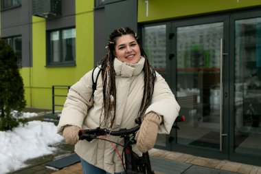 Rasta bukleli bir kadın enerji dolu bir şekilde bisiklet sürüyor. Günlük giysiler giyiyor ve önündeki yola odaklanmış görünüyor. Kendine güvenerek direksiyon sallıyor. Parlak gün ışığı