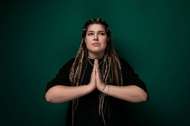 Rasta saçlı bir kadın canlı yeşil bir arka planın önünde duruyor. Kameraya tarafsız bir ifadeyle bakıyor..
