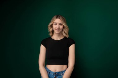 Katı yeşil bir duvarın önünde duran bir kadın. Doğrudan kameraya bakıyor gibi görünüyor, tarafsız bir yüz ifadesi sergiliyor. Duvar, olay yerine canlı bir zemin sağlıyor..