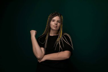 Rastalı bir kadın kollarını kavuşturup kendine güvenen bir duruş sergiliyor. Yüz ifadesi nötr ve doğrudan ileriye bakıyor..