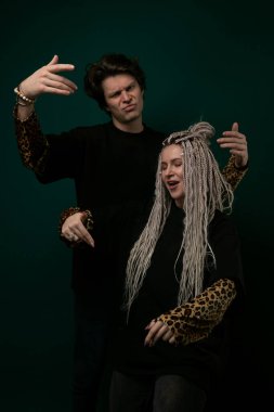 Bir kadın ve erkek kameraya gülümseyen bir pozisyonda yan yana duruyorlar. Adamın kolu kadının omzunda, rahat ve mutlu görünüyorlar..