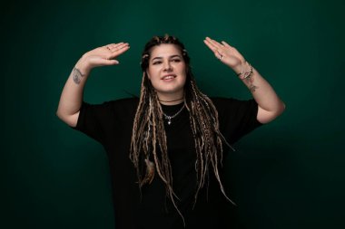 Rasta saçlı bir kadın canlı yeşil bir arka planın önünde kendinden emin bir şekilde duruyor. Saçları kalın, düğümlü rasta şeklinde ve doğrudan kameraya bakıyor..