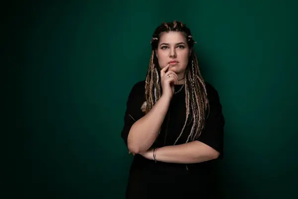 stock image A woman with dreadlocks is standing in front of a vivid green wall, looking directly at the camera. She is posing confidently with her hands by her sides, showcasing her unique hairstyle.