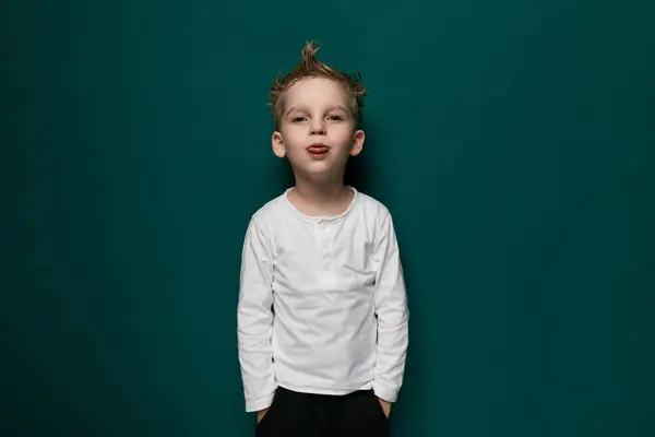 stock image A young boy with his tongue out stands in front of a bright green wall, displaying a playful expression. He seems to be enjoying himself and showing his cheeky side.