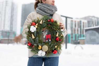 Karın üzerinde elinde geleneksel bir Noel çelengi tutan bir kadın. Karla kaplı zemin, çelengin canlı yeşiliyle çelişiyor ve şenlikli bir manzara yaratıyor..