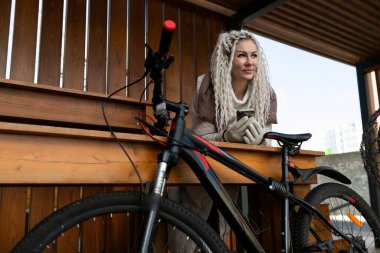 Bir kadın park edilmiş bir bisikletin yanındaki bankta oturuyor. Rahatlamış görünüyor, etrafa bakıyor. Bisiklet dik duruyor, banka yaslanıyor..