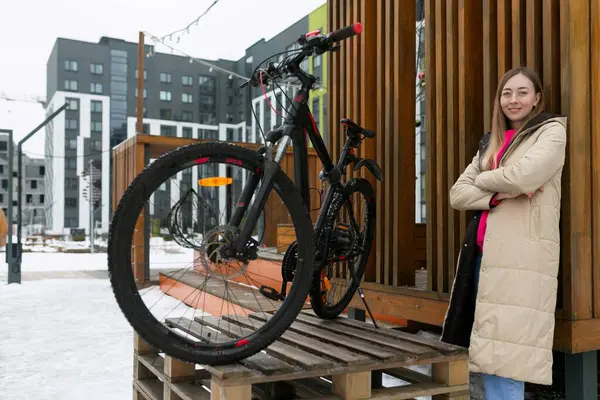 stock image A woman is standing next to a parked bike on a wooden platform. She is looking towards the distance with her hand resting on the bike. The setting is outdoors with trees in the background.