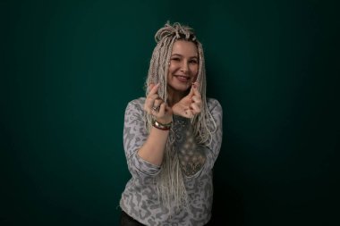 Rasta saçlı bir kadın canlı yeşil bir arka planın önünde kendinden emin bir şekilde duruyor. Benzersiz saç stili ve duruşuyla bir güç ve bireysellik hissi yayıyor..