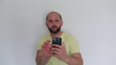 Sıradan sarı tişörtlü, sakallı, odaklanmış bir adam, akıllı bir telefonla etkileşime giriyor, tahminen beyaz bir zemine karşı durarak uygulama işlevlerini araştırıyor ya da gösteriyor..
