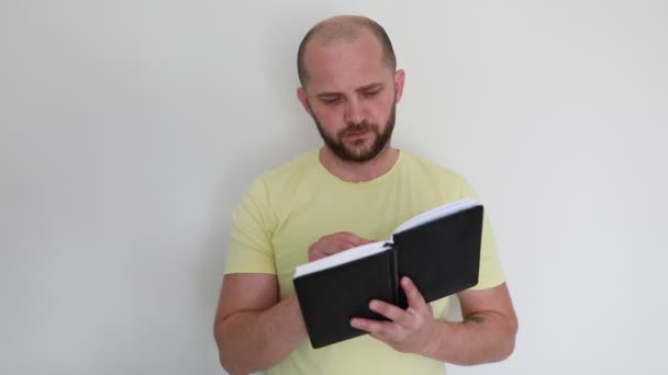 一个蓄着胡子的全神贯注的男人站在一个普通的背景下 穿着一件休闲的黄色衬衫仔细地翻阅着他拿着的黑色笔记本的书页 — 图库视频影像