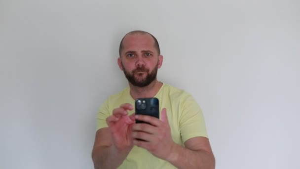 一个蓄着胡子 穿着休闲的黄色T恤的专注的男人 与智能手机互动 大概是在站在普通的白色背景下探索或演示应用程序功能 — 图库视频影像