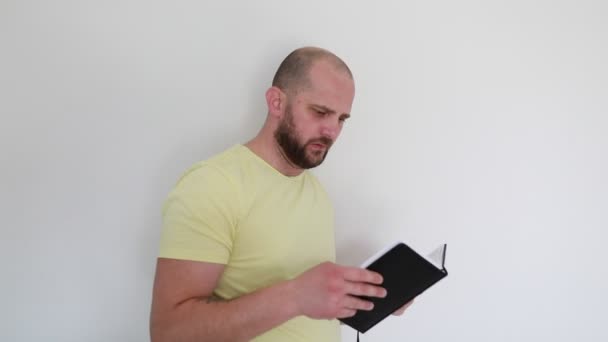一个身穿休闲黄T恤的男人站在室内 背景朴素 在翻阅他拿着的一本黑色笔记本时 露出惊讶和好奇的神色 — 图库视频影像