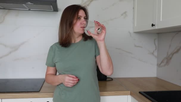 一位穿着绿色T恤衫的年轻女子 站在一个光滑的现代化厨房里 一面用大理石墙壁 一面从透明的玻璃杯里喝水 一面显得心满意足 面色苍白 — 图库视频影像