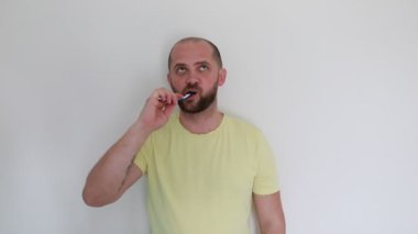 Sarı tişörtlü kısa sakallı bir adam titizlikle dişlerini fırçalayan düz beyaz bir duvara karşı duruyor. Doğal gün ışığı, rutinin sabah gerçekleştiğini gösteriyor. Ortaya çıkıyor.