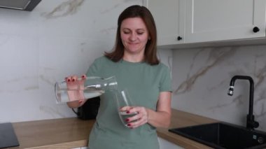 Neşeli bir kadın aydınlık, modern bir mutfakta duruyor, temiz bir bardağa kristal filtrelenmiş su döküyor. Sıradan giyinmiş ve performans sergilerken mutlu ve rahat görünüyor.