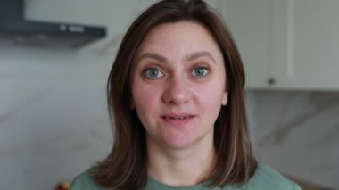 Omuz hizasında saçları ve normal bir tişörtü olan bir kadın kapalı alanda izleyiciye sıcak bir gülümseme sunarken görülüyor. Halinden memnun ve rahat görünüyor, doğal, dostane bir tavırla..