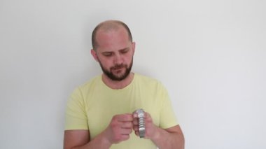 Parlak sarı tişört giyen kel bir adam parlak gümüş kol saatinin saatini titizlikle ayarlıyor. Temiz beyaz arka plan odaklandığı aktiviteyi vurguluyor..