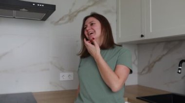 Genç bir kadın aydınlık, çağdaş bir mutfakta yüzünde ince bir gülümsemeyle duruyor. Neşeli bir aktiviteye ya da sohbete dalmış gibi görünüyor. Mutluluk hissi yayıyor.