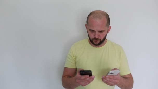 一个穿着黄色T恤的秃头男人站在一个普通的白色背景上 比较着两台智能手机 他仔细看了每一部电话 可能是评估功能或阅读内容 他的表情 — 图库视频影像