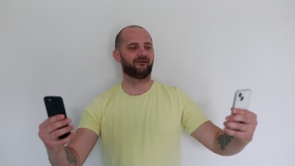Cheerful Man Stands Plain Background Holding Two Smartphones His Hands — Stock Video
