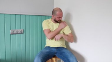 Sarı tişörtlü kel bir adam, soluk mavi bir duvara karşı ahşap bir taburede oturuyor. Omzunu rahatsız bir şekilde tutuyor, acı ya da yaralanma belirtileri gösteriyor.