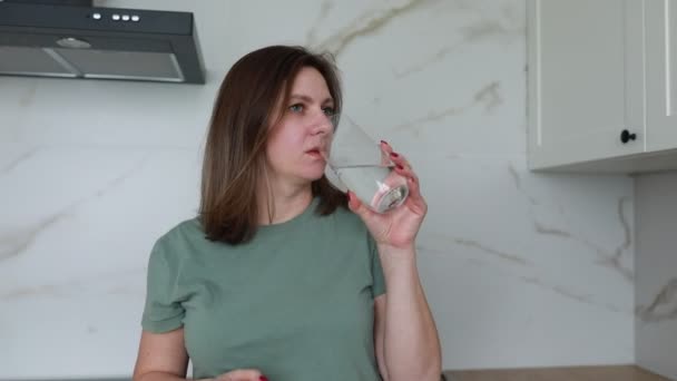 一个女人站在一个灯火通明的当代厨房里 穿着一件绿色T恤 手里拿着一个装满水的透明杯子 她似乎是在沉思 也许是在喝酒前花些时间 — 图库视频影像